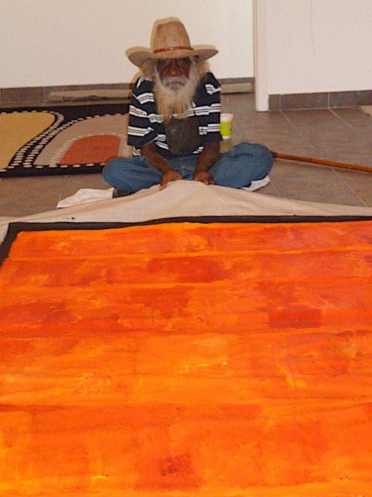 A large horizontal painting of broad washes of scarlet, orange and gold suggesting a sunlit desert horizon, with the artist seated above the canvas.