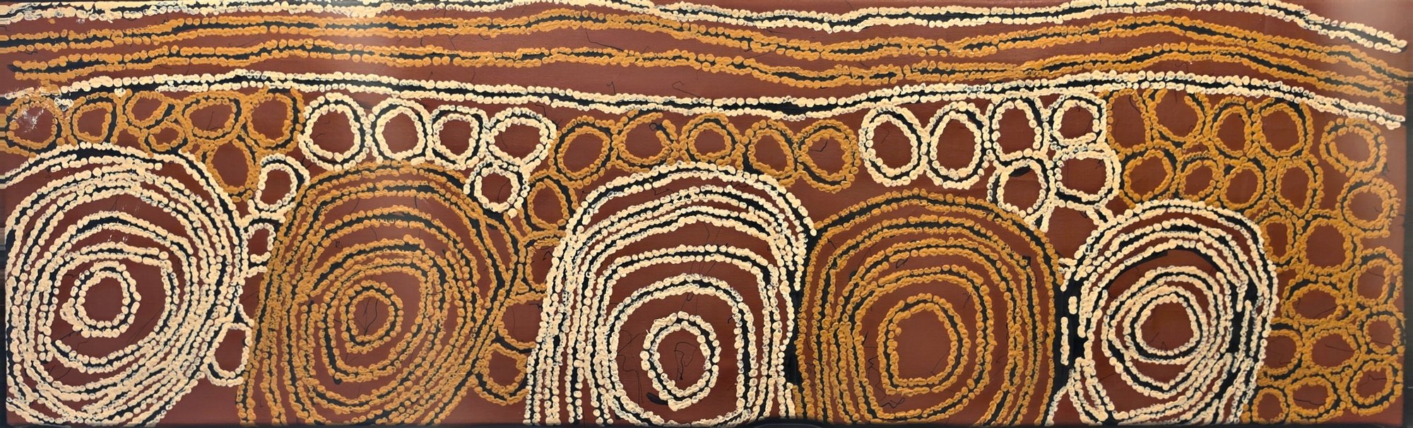Long horizontal Western Desert painting with rows of concentric circles in cream, ochre and gold on a deep red ground, suggesting sites and sandhills.
