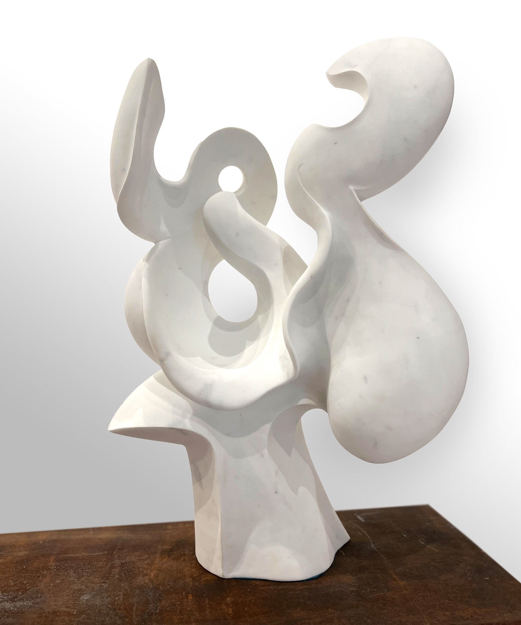 A tall white abstract sculpture with smooth flowing curves and two pierced openings, rising from a narrow base on a wooden surface.