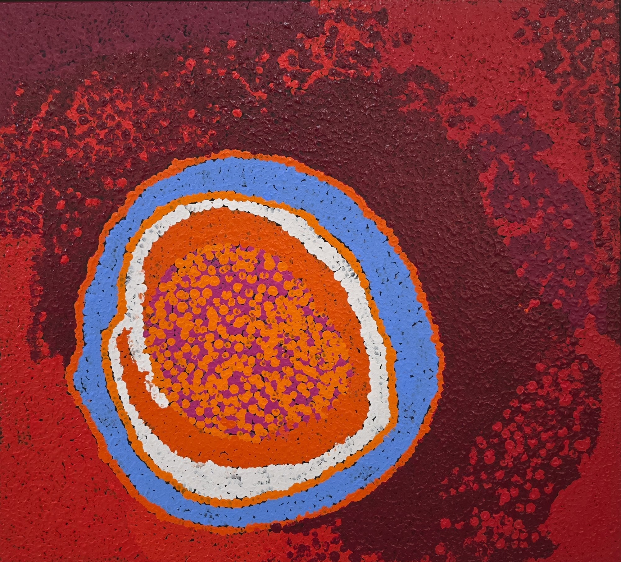 An Indigenous Australian dot painting featuring a large central oval of concentric orange, white and blue rings around a magenta and orange dotted core, set within a deep red and burgundy stippled field.